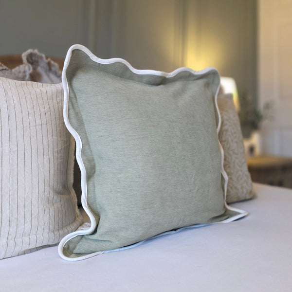 sage green scalloped edge cushion. Cream edge beading around the scalloped edge. Cushion sat on a bed with white bedding and a green wall backdrop