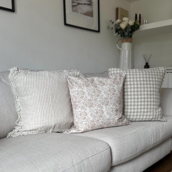 Set of 3 square cushions 1 ruffled edge linen cream with beige stripe cushion. 1 weathered edge cream and beige gingham linen cushion 1 square cream and beige floral print cotton cushion. All sat on a cream sofa in an English country inspired living room.