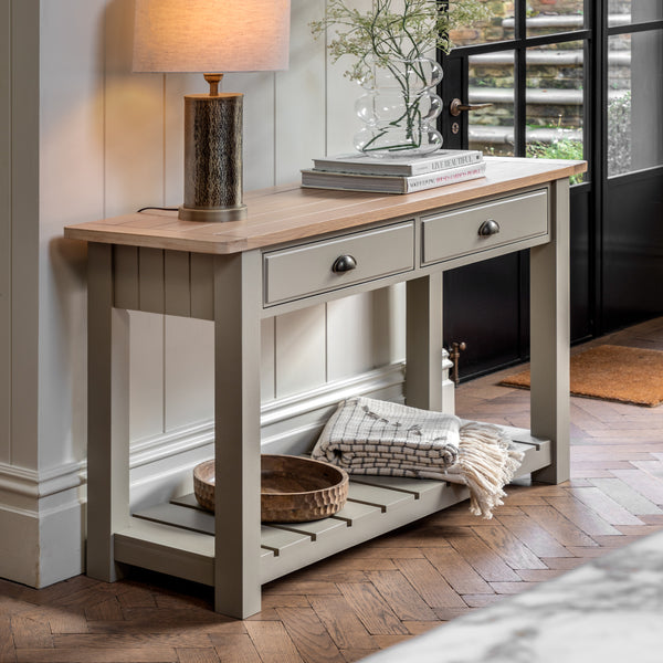 Wooden console table with 2 drawers complimented by a metal drawer pull. Grooved & planked oak top, displaying a lamp and books with a vase sat on top. A slatted bottom shelf space to store baskets, blankets and more. Sat in a light and airy hallway.