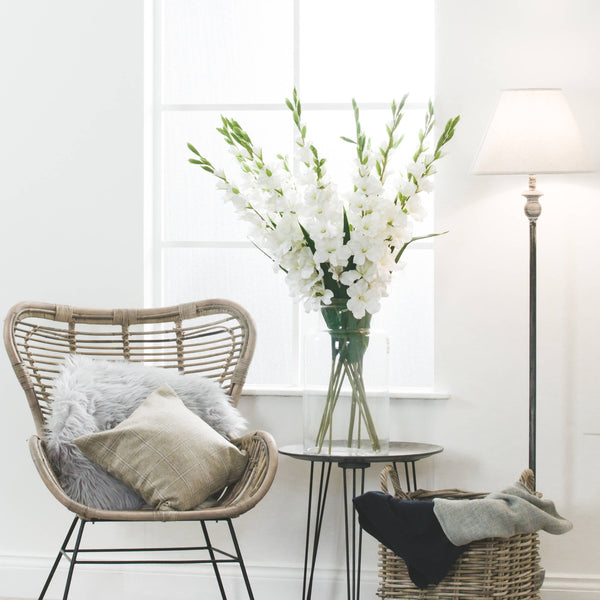 A bunch of Faux Single Stem White Gladioli in a large glass vase sat on a pin legged side table in a light and airy living room