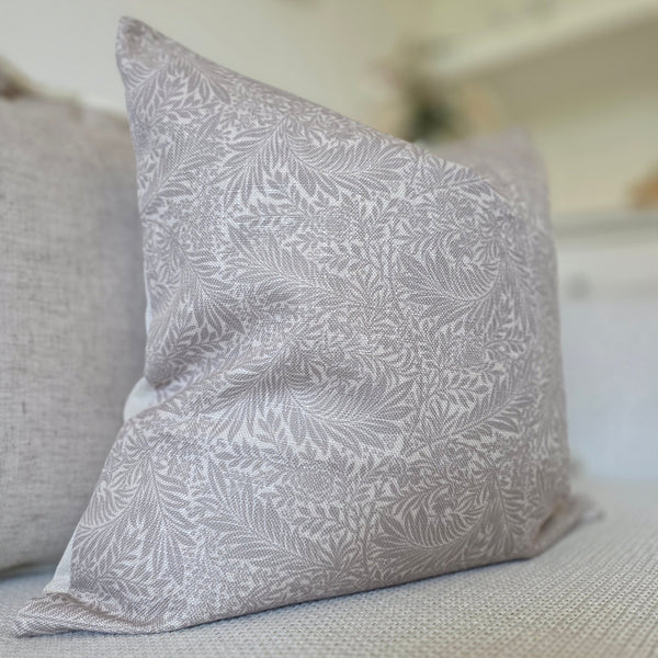 Square cushion with a greige base colour and an intricate beige leaf print. sat on a cream sofa