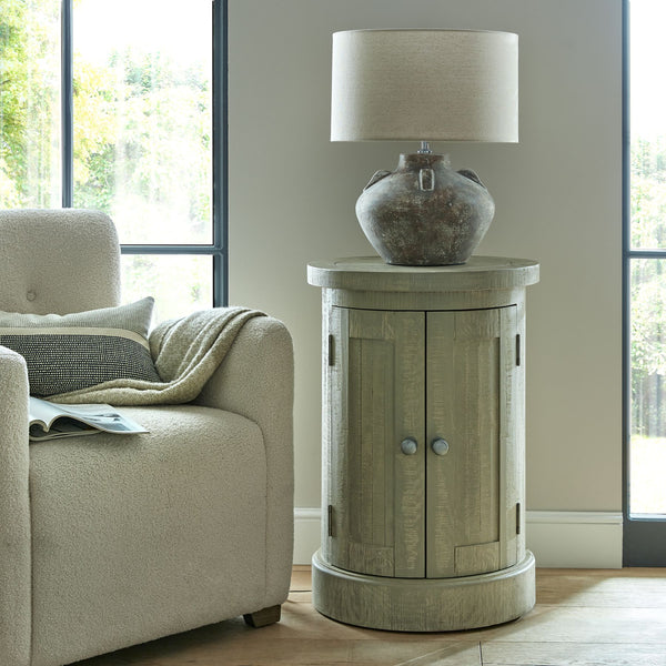 Harborview Circular Pine Wood Storage Console displaying a large brown bases and cream fabric lamp on top. Sat at the side of a cream sofa in a living room