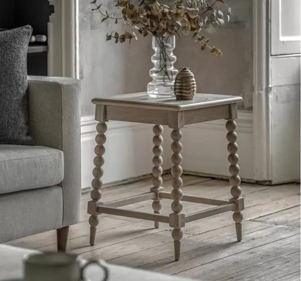 Oak Bobbin Leg Wooden Side Table. With a lime wash oak colour, square wooden top, 4 oak bobbin legs. Sat in a living room on a wooden floor. Displaying a glass vase and gold ornament