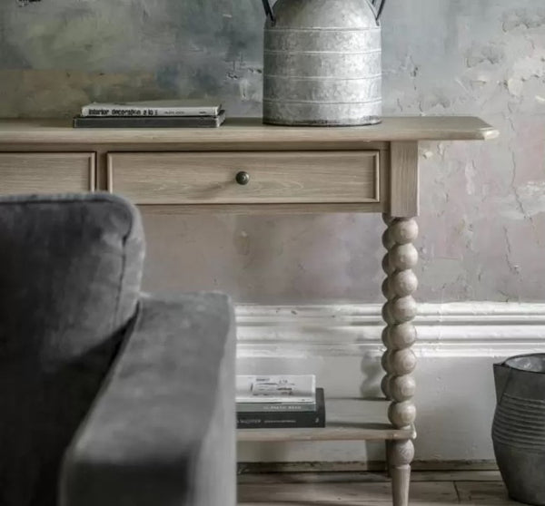 Close up of the oak Bobbin 2 Drawer Wooden rectangle top displaying a urn vase and flowers. 4 oak bobbin legs attaching a bottom shelf displaying books and a vase. 2 top rectangle drawers with a small round handle each. Sat in a moody living room