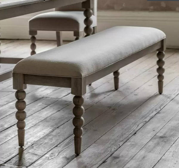 Oak Bobbin Wooden Dining Bench. Showing the biscuit coloured fabric top and beautiful oak bobbin style legs. Sat on a wooden floor in a dining room.