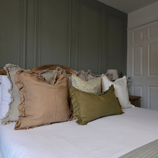 Set of 4 cushions. 1 square brown linen cushion with ruffled edge. 1 square beige cushion with an all over green leaf print. 1 square cream cushion with a repeating slim beige vertical stripe with a flanged edge. 1 rectangle olive green linen cushion with a ruffled edge. All sat on a bed with white bedding and a panelled olive green back wall.