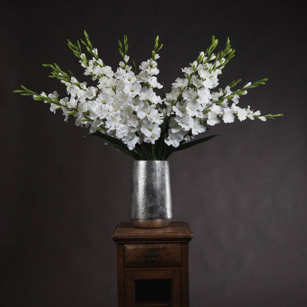 A bunch of Faux Single Stem White Gladioli in a large glass vase sat on a wooden side table in a light and dark moody room