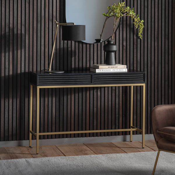 black and gold console table. rectangle black wooden slatted top with a gold metal frame. Displaying a black and gold table lamp and a black vase with green foliage.