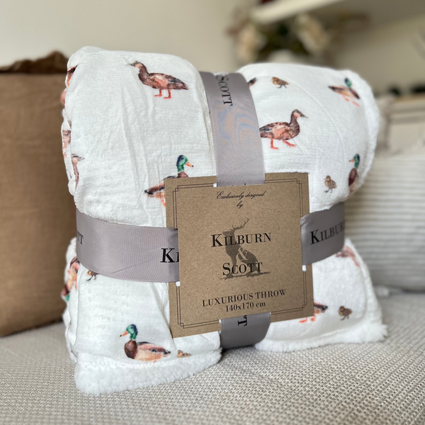 Watercolour Sherpa Duck Print Blanket. White soft and fluffy background with all over duck and duckling watercolour print. sat on a cream sofa