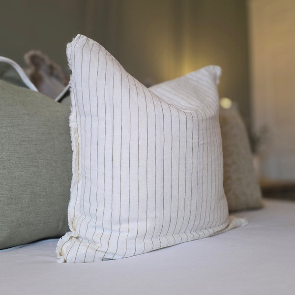 Weathered Edge Cream & Olive Linen Cushion. Sat in front of green cushions on a bed with white bedding and an olive green back wall.