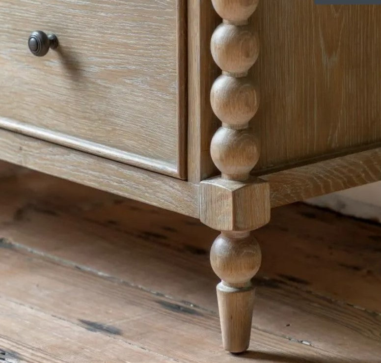 Close up of the oak bobbin legs on the 7 drawer oak bobbin chest. With a soft natural wood grain and subtle lime wash. Stood on natural real wood flooring in a bedroom