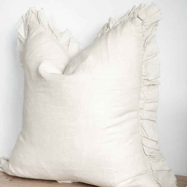 ruffled edge square cream linen cushion 45x45cm. With a frilled edge, Sat on a wooden bench against a white wall for contrast