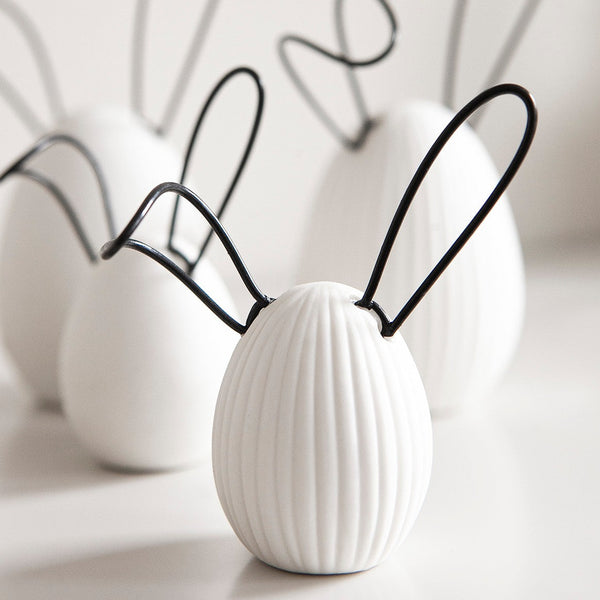 A decorative white egg with a ribbed design and black wire ears, adding a playful touch to any space. The unique design and color scheme make it a versatile addition to any decor style, perfect for Easter or springtime. Ideal for display on shelves, mantels, or desks, and makes a great gift for friends, family, or co-workers.